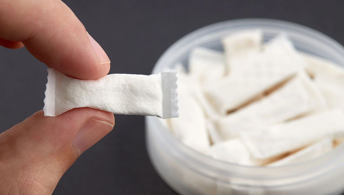Hand holding a single white nicotine pouch above an open can of pouches